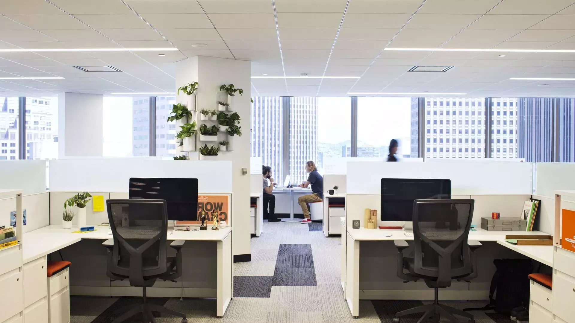 Escena interior de un edificio de oficinas en San Francisco, con escritorios en primer plano y dos personas trabajando en una mesa frente a grandes ventanales.