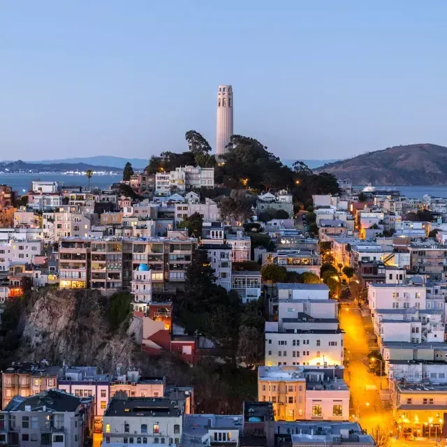 夕暮れ時のサンフランシスコのCoit Tower 。その前には明かりの灯った通り、背後にはサンフランシスコ湾が広がっています。